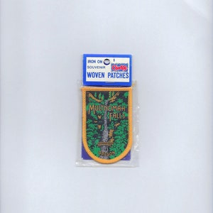 May include: Iron-on woven patch featuring a green and black image of Multnomah Falls in Oregon. The patch is surrounded by a yellow border and is packaged in a clear plastic bag with a blue and white label that reads "Iron On Souvenir Woven Patches".