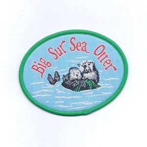 May include: A green and blue oval embroidered patch with the text "Big Sur Sea Otter" in red and white lettering. The patch features a cartoon image of two sea otters swimming in the ocean.