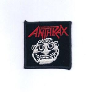 May include: A black square patch with the red word "ANTHRAX" above a white, cartoonish face. The face has large eyes and a wide, toothy grin. The patch is likely for a jacket or bag, representing a band.