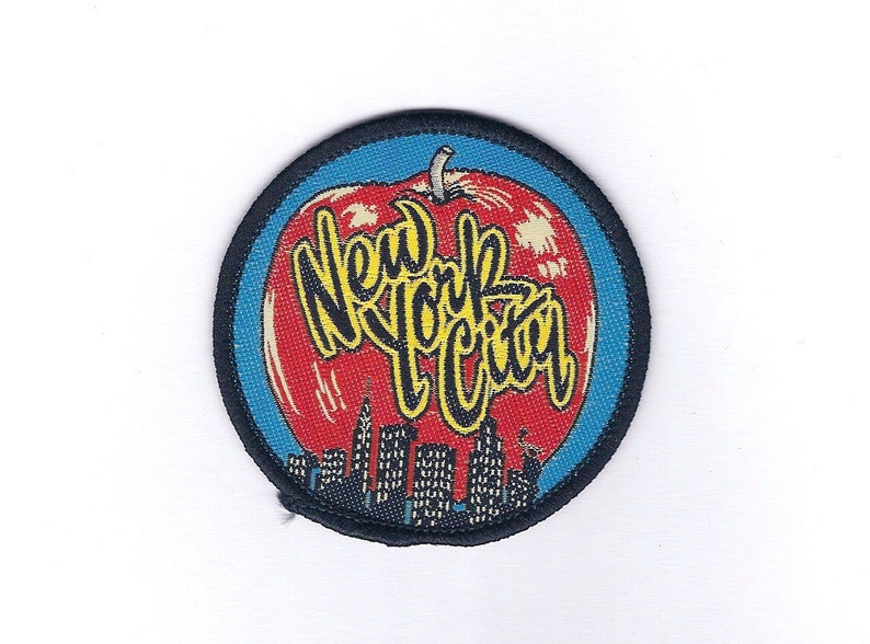 May include: A round, black-bordered patch with a red apple design. The apple has a yellow outline and the words "New York City" written in yellow letters.