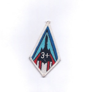 May include: A diamond-shaped patch with a blue, white, and red design. A black jet fighter is in the center of the patch with the number "3+" in white.