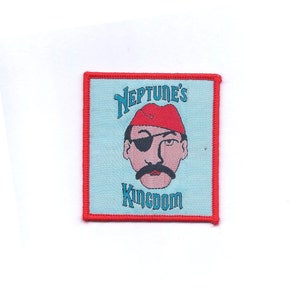 May include: Square patch with a light blue background and a red border. The patch features a cartoon pirate with a red hat, eye patch, and mustache. The text "NEPTUNE'S KINGDOM" is arched above and below the pirate's face in blue.