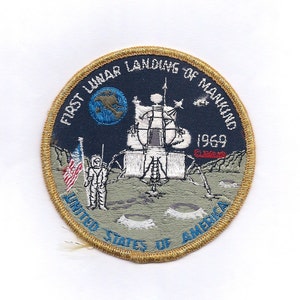 Peut inclure: Un écusson brodé doré avec le texte "First Lunar Landing of Mankind 1969" et une représentation du module lunaire atterrissant sur la lune. L'écusson comprend également le texte "United States of America".