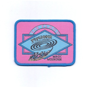 May include: A pink and blue embroidered patch with the text "Six Flags Magic Mountain" and "Cyclone The Ultimate Mindbender". The patch features a cartoon image of a roller coaster.