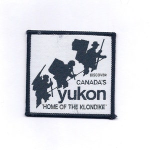 May include: A white and black embroidered patch with a silhouette of three people carrying backpacks and walking on a trail. The text "Discover Canada's Yukon" and "Home of the Klondike" is printed on the patch.