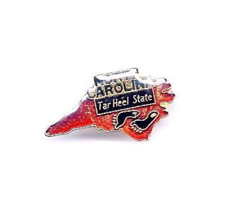 May include: A gold-colored pin shaped like the state of North Carolina. The pin is red and orange with a black banner that says "Tar Heel State" and "NORTH CAROLINA" in gold letters.