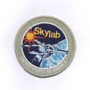 May include: A circular embroidered patch with a grey border and a blue background. The patch features a space station orbiting Earth with a sun in the background. The text "Skylab" is in yellow lettering above the space station. The text "United States of America" is in white lettering around the bottom of the patch.