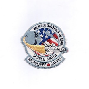 May include: A blue and white embroidered patch with the names of the Challenger astronauts: Mcnair, Onizuka, Resnik, Scobee, Smith, McAuliffe, and Jarvis. The patch features a space shuttle flying over a globe with the American flag in the background.