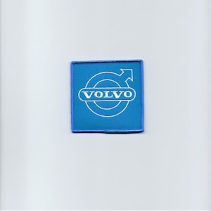 May include: A blue fabric patch with a white embroidered Volvo logo. The logo features a circle with an arrow pointing to the right and the word "VOLVO" written inside the circle.