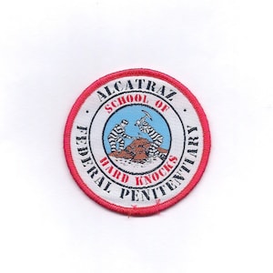May include: A red, white, and blue embroidered patch with the text "Alcatraz School of Hard Knocks Federal Penitentiary". Two cartoon prisoners are shown digging in the ground with picks.