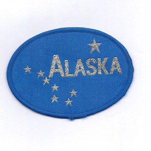 Peut inclure: Patch ovale bleu avec le mot "ALASKA" en broderie argentée et une étoile argentée dans le coin supérieur droit. Il y a sept petites étoiles argentées autour du mot "ALASKA".