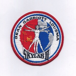 May include: A white and red circular patch with blue trim. The patch features a white figure with outstretched arms, a red and blue globe, and the text "BEAN GARRIOTT LOUSMA SKYLAB II".