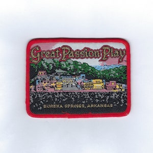 May include: A red, white, and gold embroidered patch with the text "Great Passion Play" and "Eureka Springs, Arkansas". The patch features a colorful illustration of a building with a mountain in the background.