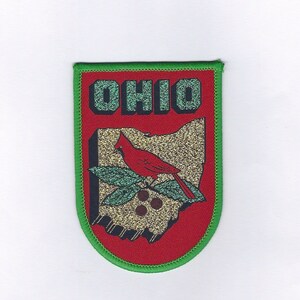 May include: A red, green, and gold embroidered patch with the word "OHIO" in large letters and a red cardinal bird perched on a branch with berries. The patch is shaped like the state of Ohio.