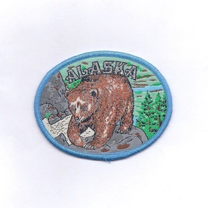 May include: A blue and green embroidered patch with a brown bear and the word "ALASKA" in white lettering.