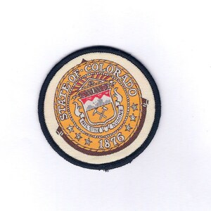 May include: A round, embroidered patch with a black border. The patch features the state seal of Colorado, which includes a shield with a mountain range, crossed mining picks, and the words "Nil Sine Numine" and "1876". The seal is surrounded by the words "State of Colorado" and five stars.