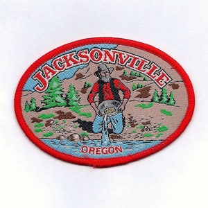 May include: An embroidered patch with the text "JACKSONVILLE OREGON" and a cartoon image of a man panning for gold in a river. The patch is red, white and blue.