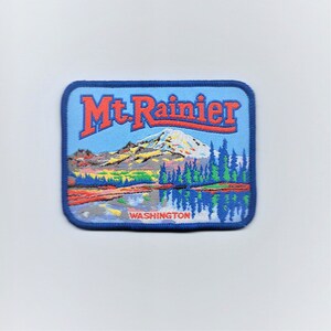May include: A blue and red embroidered patch with the text "Mt. Rainier" and a depiction of a mountain with a lake in front of it. The text "WASHINGTON" is below the image.