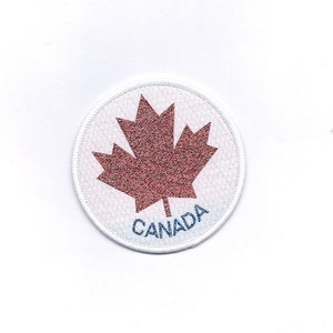 May include: A round patch featuring a red maple leaf, a national symbol of Canada, on a white background. The word "CANADA" is printed in blue below the leaf. The patch has a white stitched border.