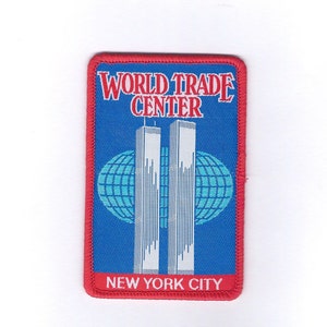 Peut inclure: Un écusson brodé rouge, blanc et bleu avec les mots "World Trade Center" et "New York City". L'écusson représente une image stylisée des tours jumelles avec un globe derrière elles.