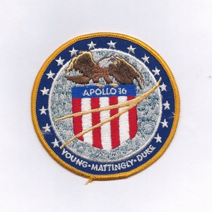 May include: A blue and gold embroidered patch with a white eagle, a red, white, and blue shield with the text "APOLLO 16", and the text "YOUNG - MATTINGLY - DUKE" around the edge.