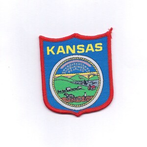 May include: A blue and red embroidered patch with the word "KANSAS" in yellow letters and the state seal of Kansas in the center.