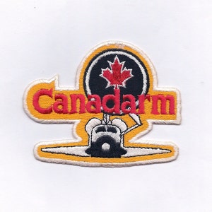 May include: A yellow and white embroidered patch with the text "Canadarm" in red lettering. A black circle with a red maple leaf is above the text. A black and white illustration of a robotic arm is below the text.