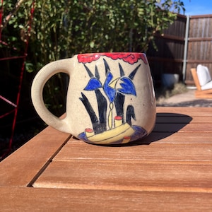 May include: A beige ceramic mug with a curved handle. The mug features a hand-painted design of blue flowers, black stems, and a yellow boat with red accents. The rim is decorated with a red and white pattern.