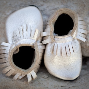 May include: A pair of silver leather baby moccasins with fringe trim.