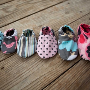 May include: Six pairs of baby shoes in various colours and patterns, including polka dots, stripes, and animal prints. The shoes are laid out on a wooden surface.