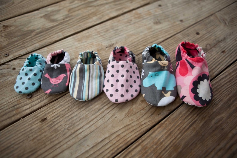 May include: Six pairs of baby shoes in various colours and patterns, including polka dots, stripes, and animal prints. The shoes are laid out on a wooden surface.