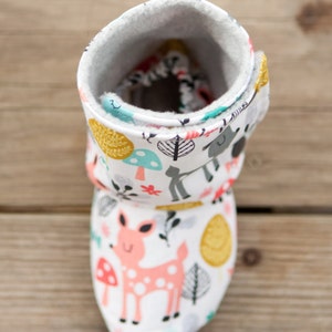 May include: White baby booties with a woodland animal print. The booties feature a pink deer, mushrooms, and leaves. The booties are lined with a soft, white fleece.