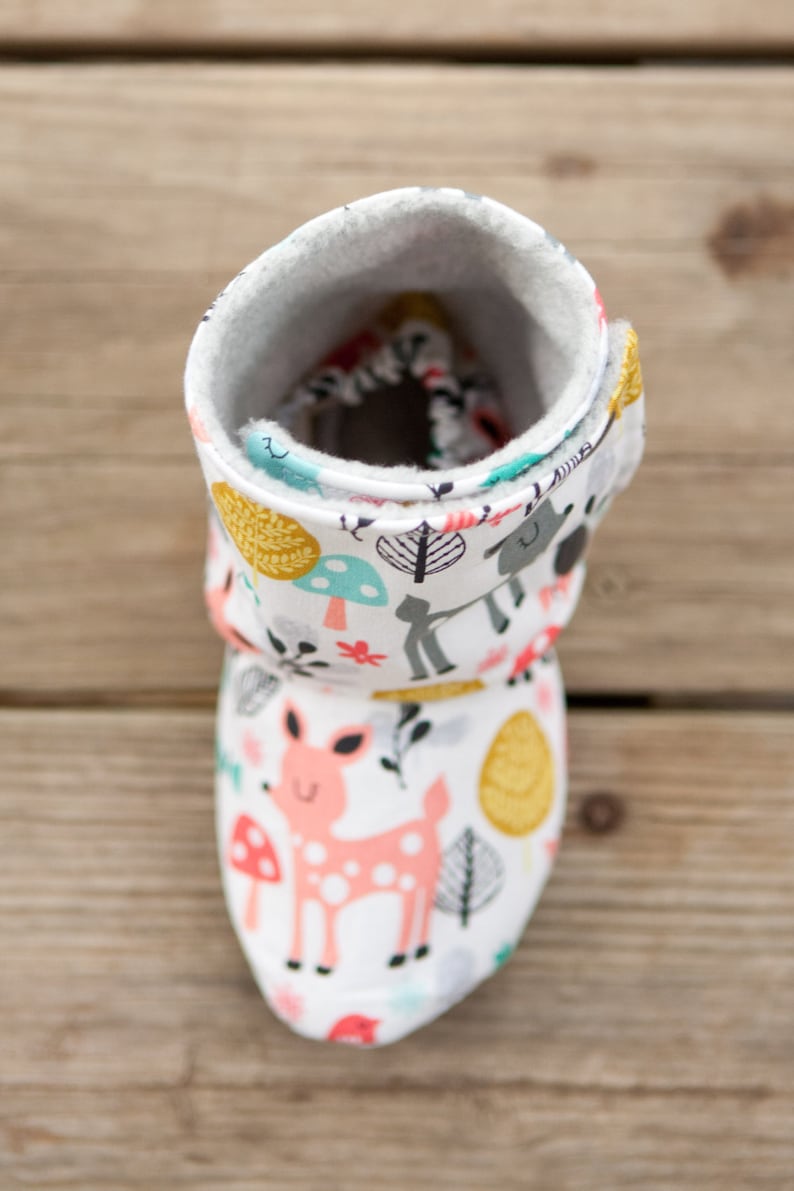 May include: White baby booties with a woodland animal print. The booties feature a pink deer, mushrooms, and leaves. The booties are lined with a soft, white fleece.