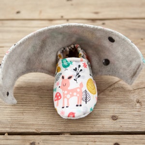 May include: A pair of white baby booties with a woodland animal print featuring a pink deer, green birds, and yellow leaves. The booties are lined with gray fabric and have black buttons.