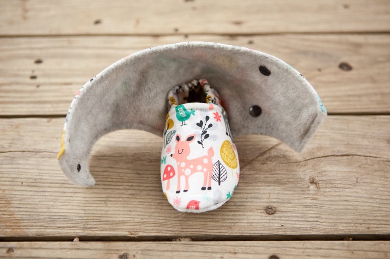 May include: A pair of white baby booties with a woodland animal print featuring a pink deer, green birds, and yellow leaves. The booties are lined with gray fabric and have black buttons.