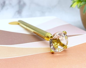 May include: A gold pen with a large clear crystal on the end. The pen is lying on a pink and white notebook.