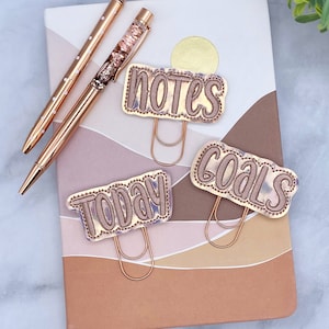 May include: Three gold-toned paper clips with decorative fabric covers. The covers have the words "notes", "goals", and "today" written on them. The clips are on a notebook with a brown and pink design.