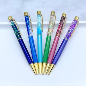 May include: Seven glitter pens with gold caps and different colored bodies. The pens are arranged in a fan shape on a white surface.