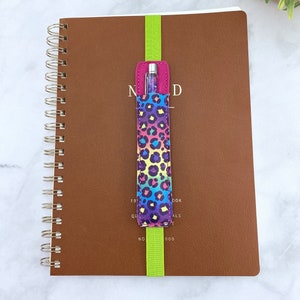 May include: A brown leather bound notebook with a green elastic bookmark and a rainbow leopard print pen holder with a silver pen inside.