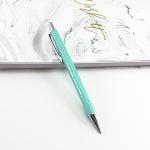 May include: A turquoise glitter pen with a silver tip and a silver cap. The pen is lying on a white surface.