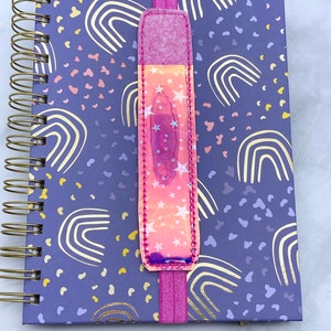 May include: A purple notebook with a gold rainbow pattern and a pink elastic bookmark with a holographic star design.