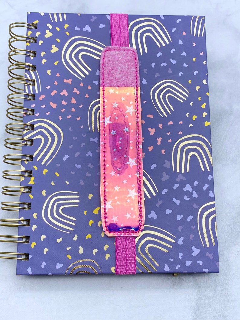 May include: A purple notebook with a gold rainbow pattern and a pink elastic bookmark with a holographic star design.