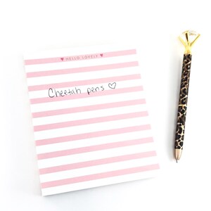 May include: A pink and white striped notepad with the text "Hello Lovely" and "Cheetah pens" written on it. A gold pen with a diamond-shaped top and a cheetah print design is next to the notepad.