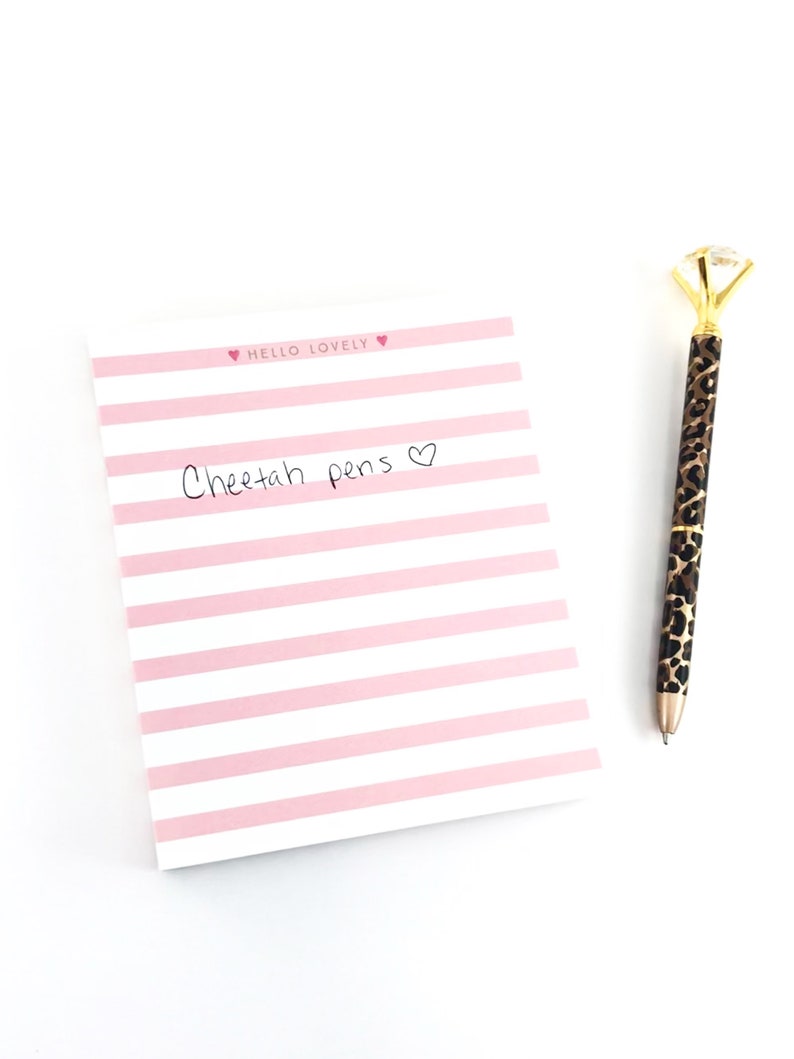 May include: A pink and white striped notepad with the text "Hello Lovely" and "Cheetah pens" written on it. A gold pen with a diamond-shaped top and a cheetah print design is next to the notepad.