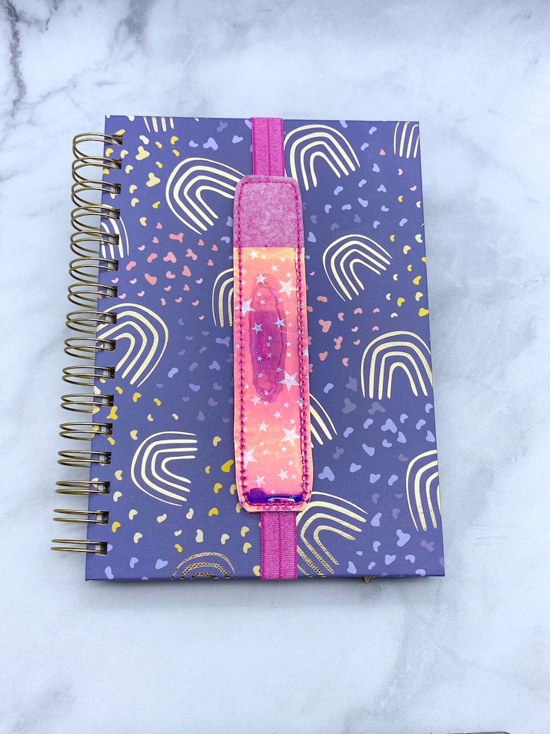 May include: A purple notebook with gold foil rainbow designs and a pink elastic bookmark with iridescent stars.