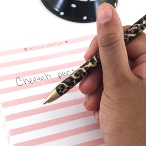 May include: A close-up of a hand holding a cheetah print pen. The pen is being used to write the words "Cheetah pens" on a pink and white striped notepad with the words "Hello Lovely" written on the top.