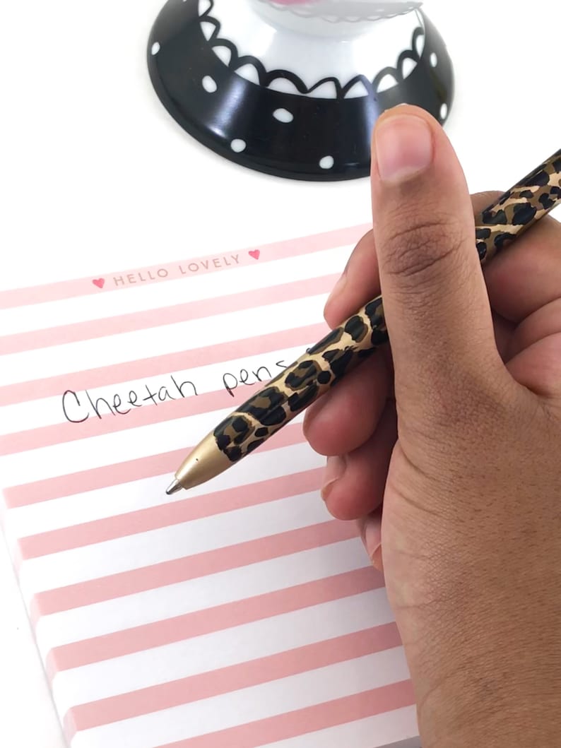 May include: A close-up of a hand holding a cheetah print pen. The pen is being used to write the words "Cheetah pens" on a pink and white striped notepad with the words "Hello Lovely" written on the top.