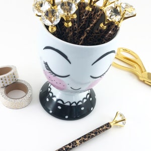 May include: A white ceramic mug with a cartoon face and black polka dots, filled with brown and gold pens with clear rhinestone tops.