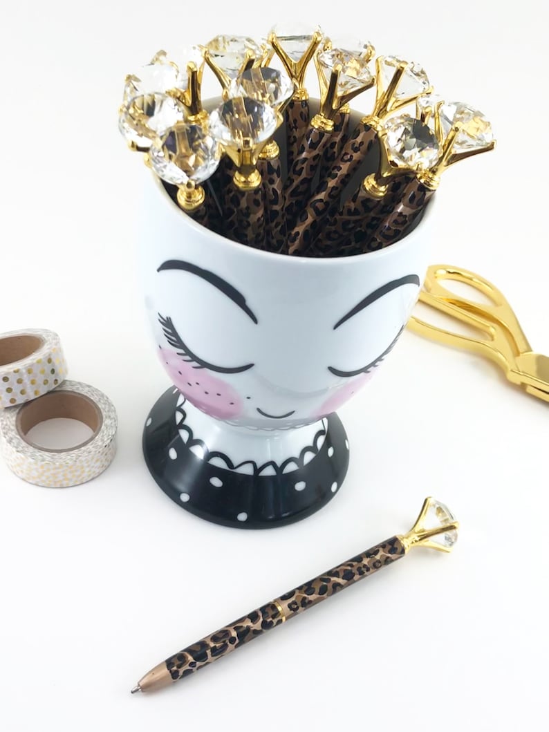 May include: A white ceramic mug with a cartoon face and black polka dots, filled with brown and gold pens with clear rhinestone tops.