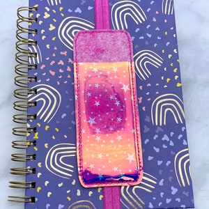 May include: A purple notebook with a gold spiral binding and a pink and iridescent star-patterned bookmark. The notebook has a rainbow and polka dot pattern.